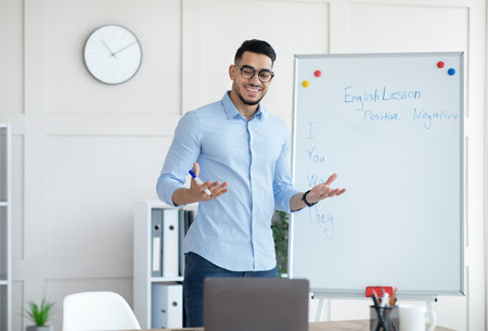 Cheerful young Arab teacher standing near blackboard and explaining English grammar to students online from homeの写真素材