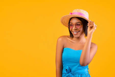 Summer vibes. Portrait of cheerful young black lady in dress, straw hat and sunglasses looking at...