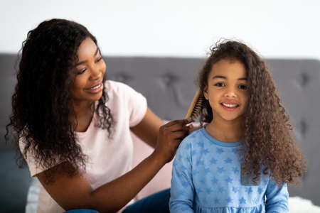 Beautiful black woman brushing her daighters hair on bed indoorsの写真素材