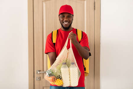 Black deliveryman holding eco mesh bag with groceriesの写真素材