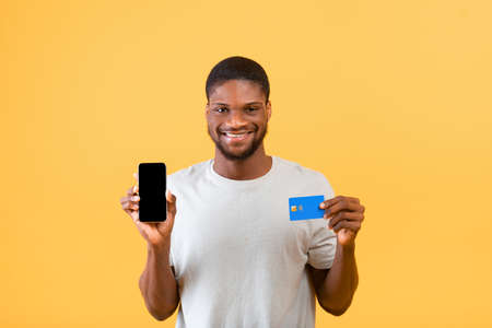 Excited black guy holding phone with empty screen and credit card on yellow background, promoting...