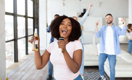 Millennial black lady with beer bottle and mic singing karaoke for her diverse friends at home