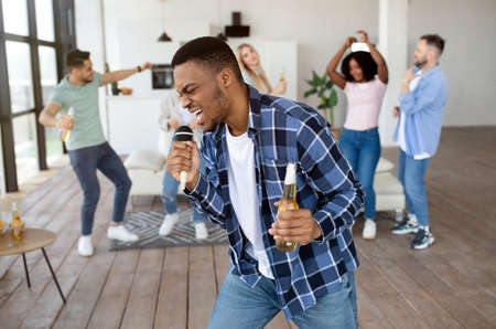 Emotional black guy with bottle of beer singing karaoke on student party with his international friends at homeの写真素材