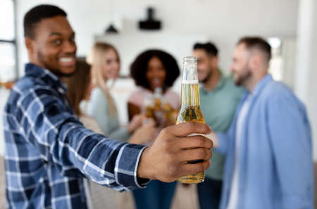 Funky black guy showing bottle of beer, celebrating with group of his diverse friends, saying CHEERS...