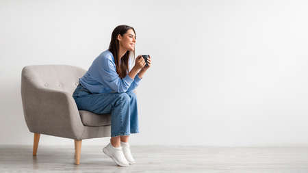 Full length of young woman having coffee break in cozy armchair against white studio wall, banner...