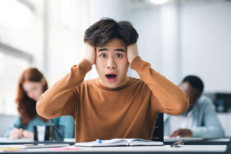 Asian male student grabbing head sitting at desk in classの素材 [FY310172485915]