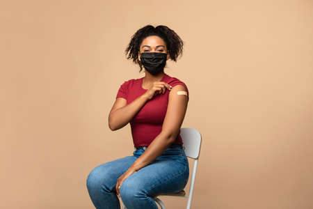 Vaccinated black woman in face mask showing arm with plaster aid, sitting with rolled up sleeve after covid-19 vaccine shotの写真素材