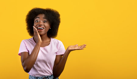 Wow, cool offer. Excited black woman holding hand, showing free space on palm and touching cheek, yellow backgroundの写真素材