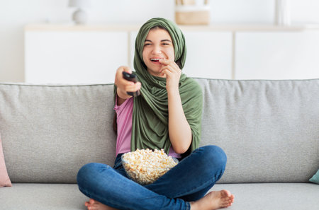 Home entertainments. Full length of Muslim teenage girl in hijab with remote control eating popcorn and watching TVの写真素材