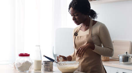Young african american lady wearing apron cracking egg and adding to other ingredients, preparing...