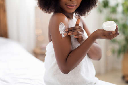 Unrecognizable black lady applying body cream on shoulder after shower in morning at homeの写真素材