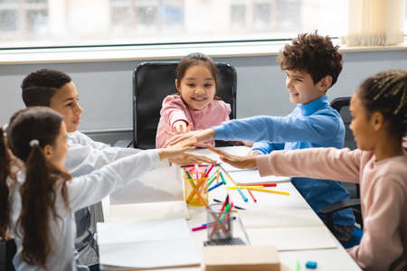 Happy multiethnic schoolchildren putting hands together at classroom
