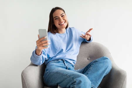 Cheerful young woman having online business meeting on smartphone, sitting in cozy armchair on white...