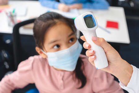 Teacher with contactless thermometer measuring temperature of asian pupil