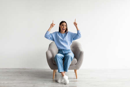 Excited Caucasian woman sitting in armchair, pointing two index fingers up at empty space above her head, mockupの写真素材