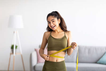 Successful weight loss. Slim Indian woman measuring waist with tape, satisfied with slimming program at homeの写真素材