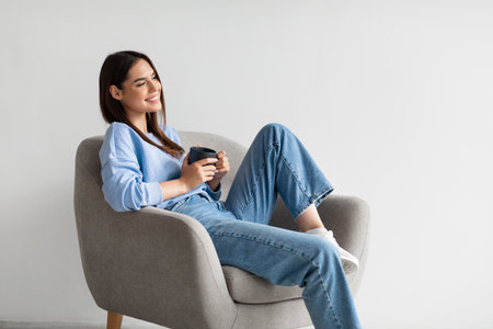 Portrait of happy young woman sitting in armchair with cup of hot coffee, relaxing on white studio backgroundの写真素材