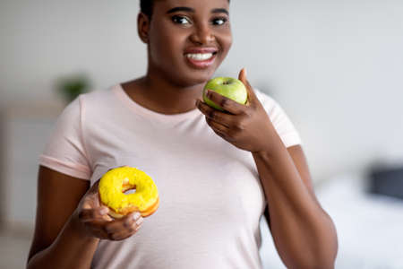 Plump Afro woman choosing between apple and donut, selecting fruit, preferring healthier nutrition for weight lossの写真素材