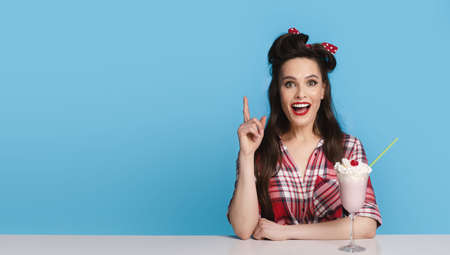 Excited pinup woman in retro clothes sitting at table with milk shake, gesturing eureka over blue background, copy spaceの写真素材