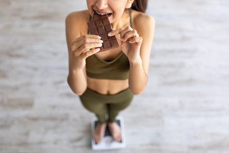 Diet failure, cheat meal concept. Above view of Indian woman eating chocolate while standing on scalesの写真素材