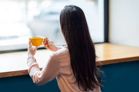 Unrecognizable young woman sitting in cafe and looking to outside the window, enjoying morning coffeeの写真素材