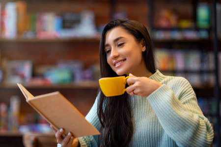 Young arab lady reading favourite book and enjoying coffee in cafe, sitting in cafeteria and spending time with pleasureの写真素材