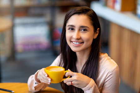 Coffee time. Portrait of happy arab woman with mug of hot beverage, resting in cafe and looking at camera, free spaceの写真素材