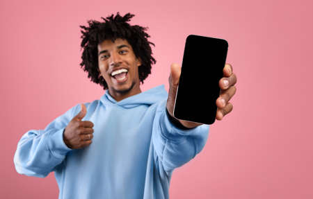 Joyful Afro teen guy showing smartphone with mockup for website or app design, gesturing thumb up, pink backgroundの写真素材