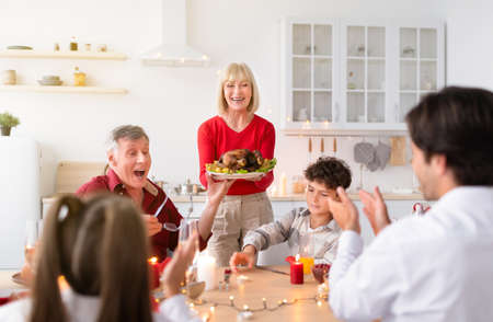 Senior woman serving traditional holiday turkey for multi generation family, celebrating...