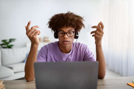 Serious Afro teenager using laptop and headphones to communicate with his university teacher or...