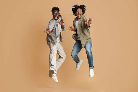 Two Cheerful Emotional African American Guys Jumping Over Beige Studio Background