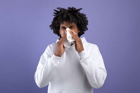 Cold and flu. Sick black teenager sneezing in paper tissue, suffering from rhinitis or sinusitis on violet backgroundの写真素材