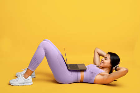Online gym. Fit african american lady doing abs exercise sit-ups, training with laptop, exercising on yellow backgroundの写真素材