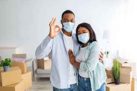 Portrait of loving african american couple in masks showing keys, standing in own house full of carton boxesの写真素材