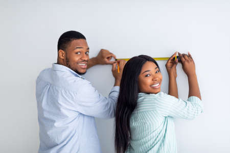 Happy black couple measuring wall with tape-measure, doing repairs in new apartment togetherの写真素材