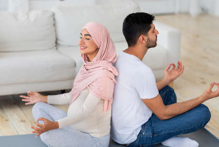 Yoga for pregnancy. Happy muslim spouses meditating together at home, sitting back to back and practicing lotus poseの写真素材