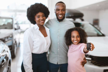 Cute black girl showing car key, standing with her parents at auto dealership, selective focus. Copy spaceの写真素材