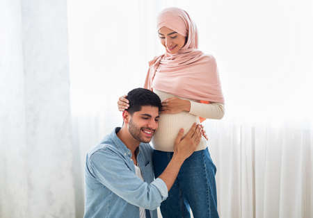 Happy arab man pressing his ear against his wifes pregnant belly, listening to his babys heartbeat, free spaceの写真素材