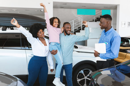 Black family feeling excited over buying auto, raising hands, gesturing YES, standing with car salesman at dealershipの写真素材