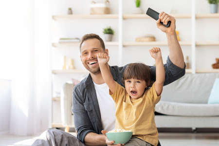 Positive father watching football game on tv with his little son, emotional boy shouting enjoying...