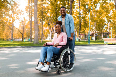 Full length of cheerful black guy taking his impaired girlfriend in wheelchair for walk at city park...