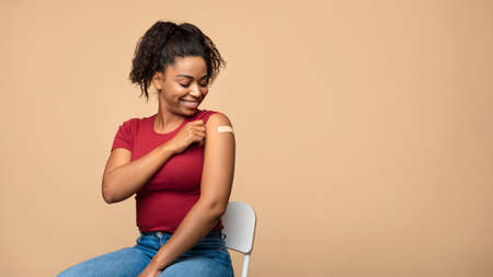 Cheerful vaccinated Afro woman showing plaster on her shoulder after immunization against covid over beige backgroundの写真素材