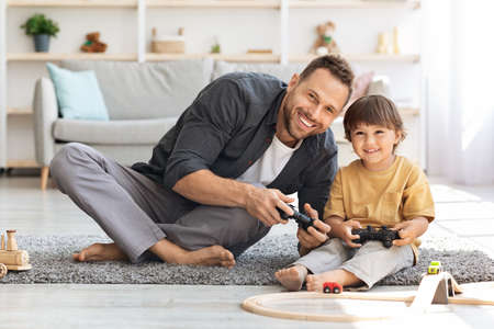 Happy young dad and his little son playing video games together, sitting on floor with joysticks,...