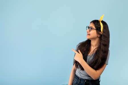 Cheery Indian teen girl pointing aside at blank space on blue studio backgroundの写真素材
