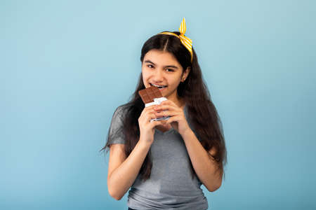 Lovely Indian teenage girl eating tasty chocolate bar, surrendering to temptation on blue studio...