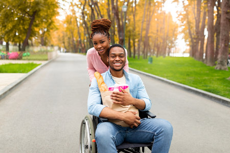 Cheerful handicapped man in wheelchair holding paper bag with food, his lovely girlfriend helping...