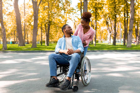 Affectionate black woman and her disabled boyfriend in wheelchair looking at each other on walk at...