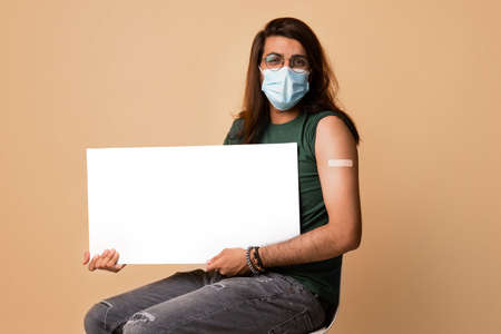 Middle-eastern young man in mask got vaccinated, holding blank boardの写真素材