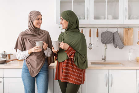 Happy Senior Muslim Mother And Daughter Drinking Coffee In Kitchenの写真素材