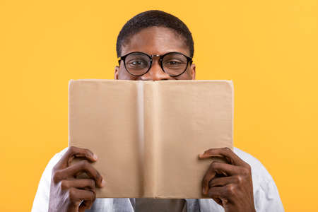 Education concept. African american guy in glasses covering face with book, hiding behind it over yellow backgroundの写真素材